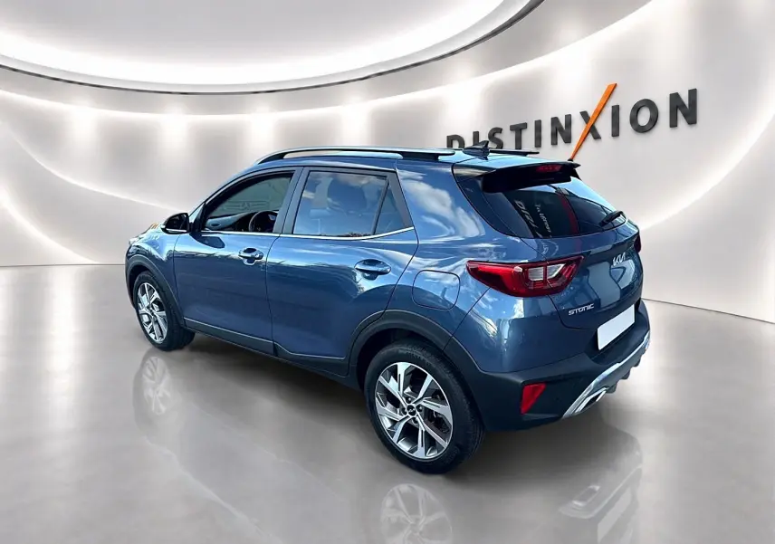KIA Stonic gris en 3/4 arrière droit, avec barres de toit et jantes alliage visibles dans un showroom lumineux.