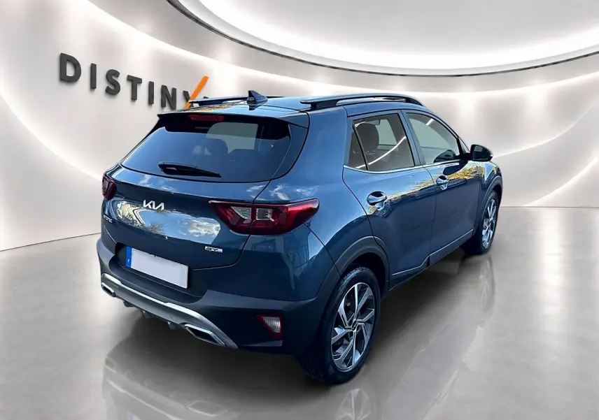 Vue 3/4 arrière droite d’un KIA Stonic gris avec jantes alu et barres de toit dans un showroom lumineux.