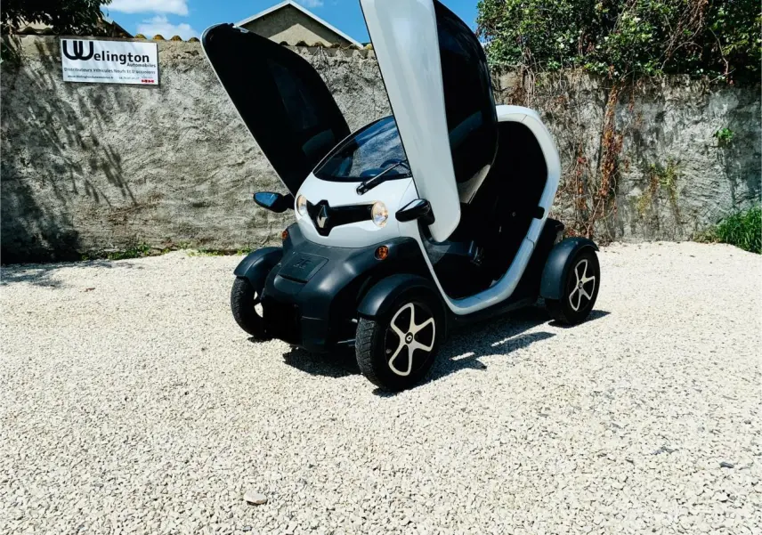 Renault Twizy blanc en 3/4 avant droit avec portes papillon ouvertes sur sol gravillonné.