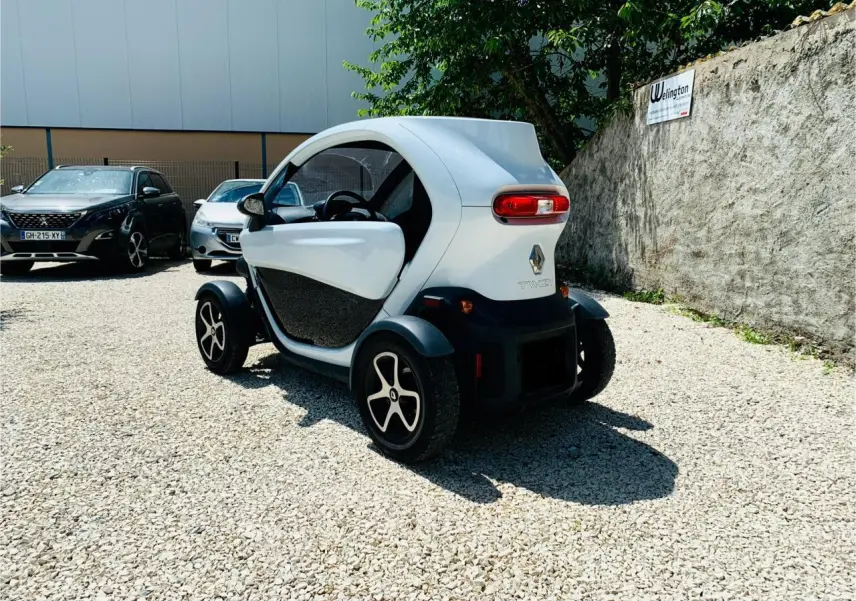Vue 3/4 arrière droite d'une Renault Twizy blanche électrique garée sur un sol gravillonné, avec portes ouvertes.