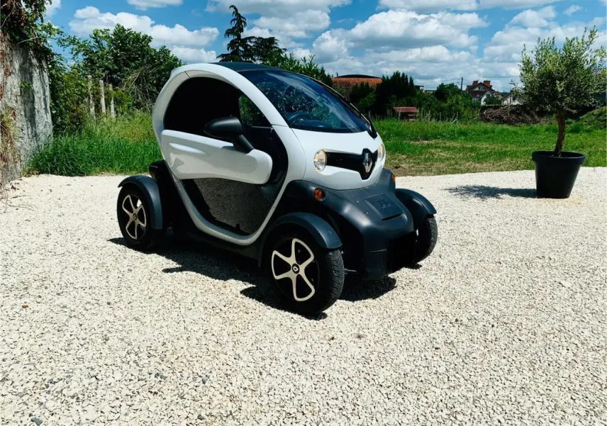 Vue 3/4 avant droite d'une Renault Twizy blanche électrique garée sur un sol gravillonné sous un ciel bleu.