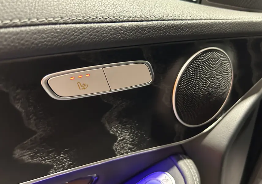 Détail intérieur côté gauche avec commande de siège chauffant active et haut-parleur rond sur panneau noir brillant.
