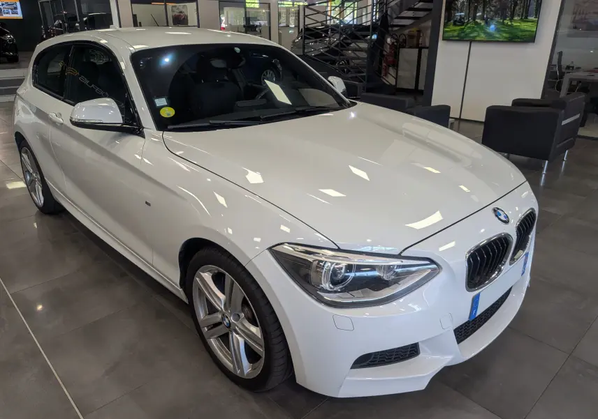 BMW Série 1 F21 118d M Sport blanc alpin vue 3/4 avant droit en intérieur avec jantes alliage 18 pouces.