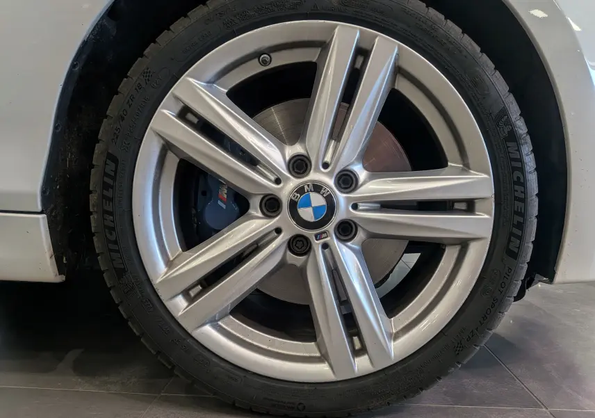Gros plan sur la roue avant gauche d'une BMW Série 1 F21 blanche, jante alliage 18 pouces avec logo M visible.
