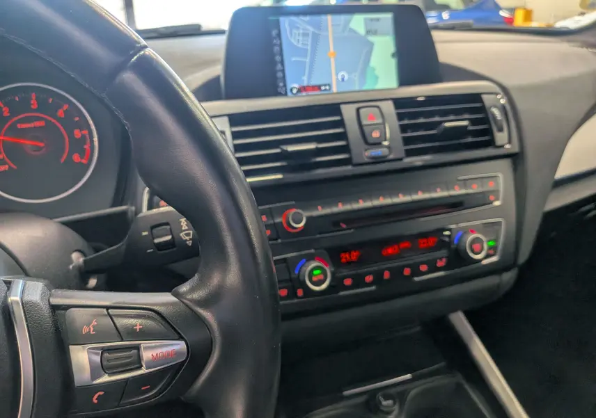 Vue intérieure centrée sur le tableau de bord et volant noir de la BMW Série 1 F21 blanc alpin avec écran GPS intégré.