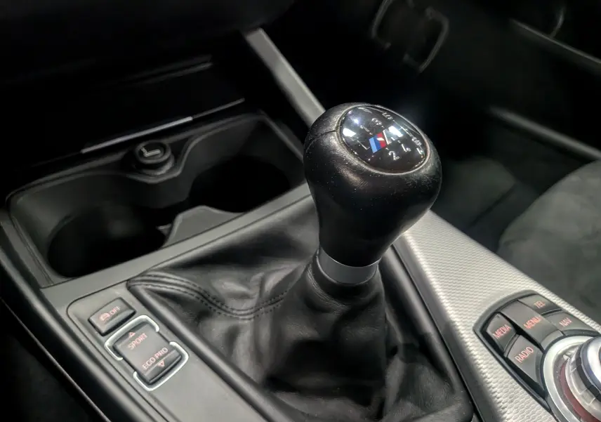 Gros plan sur le levier de vitesse manuel noir avec logo M de la BMW Série 1 F21 blanc alpin, intérieur moderne et commandes visibles.