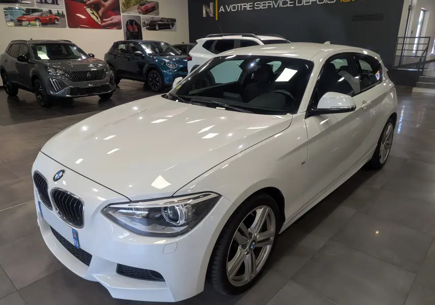 BMW Série 1 F21 118d M Sport blanc alpin vue 3/4 avant droit en intérieur avec jantes alliage 18 pouces.