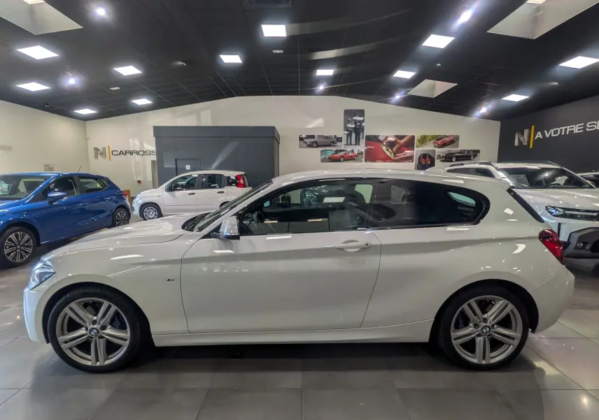 Profil latéral droit d'une BMW Série 1 F21 blanche Alpine en finition M Sport, avec jantes alliage 18 pouces, en intérieur showroom.