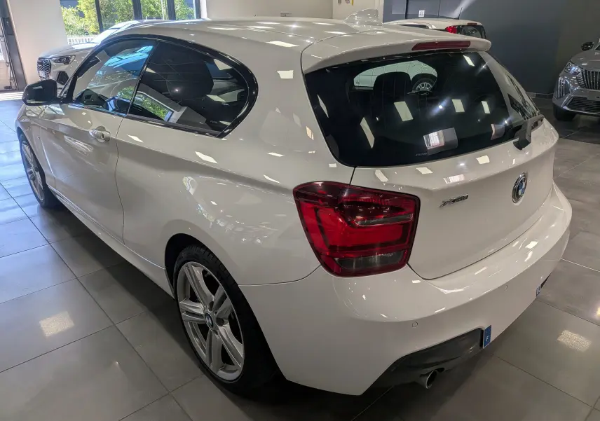 BMW Série 1 F21 blanc alpin vue 3/4 arrière droit, avec jantes alliage 18 pouces et feux arrière rouges.
