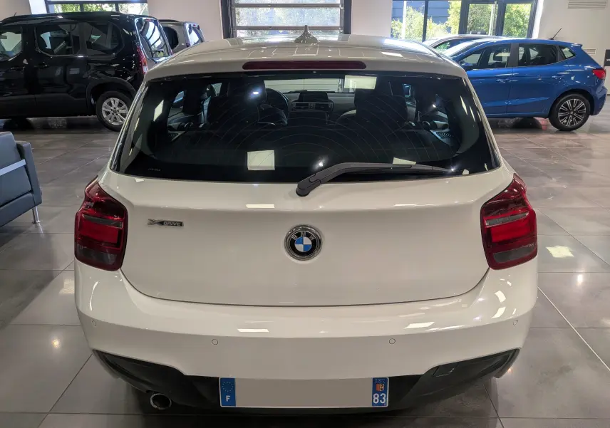 Vue arrière d'une BMW Série 1 F21 blanche Alpine avec logo BMW et badge xDrive visible en intérieur showroom.