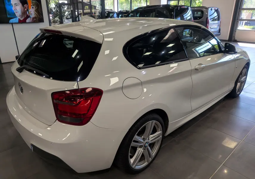 Vue 3/4 arrière droite d'une BMW Série 1 F21 118d M Sport blanche Alpine avec jantes alliage 18 pouces en showroom.