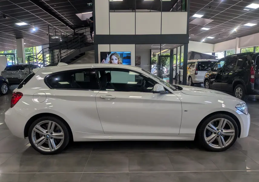 BMW Série 1 F21 blanc alpin vue de profil côté gauche en intérieur avec jantes alliage 18 pouces M Sport.
