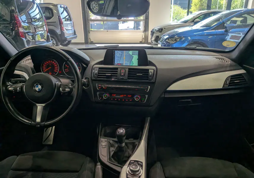 Vue intérieure avant de la BMW Série 1 F21 blanche, montrant le tableau de bord, volant, écran GPS et levier de vitesse manuel.