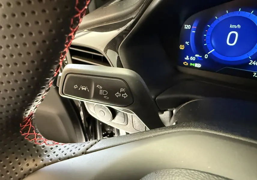 Gros plan sur la commande de clignotants et régulateur, volant noir perforé avec surpiqûres rouges, tableau de bord numérique bleu.