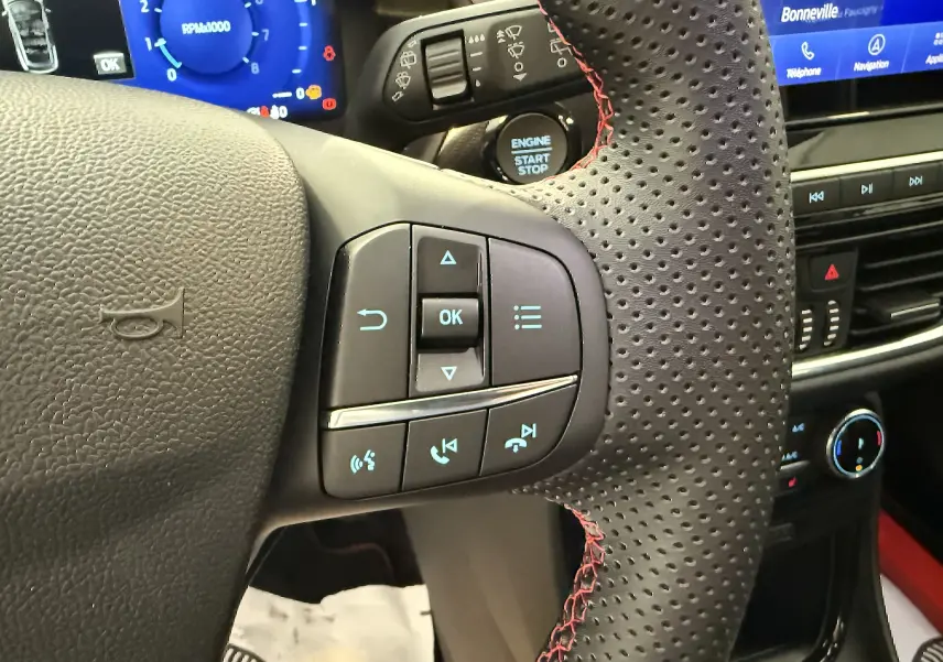 Gros plan sur le volant perforé avec surpiqûres rouges du Ford Puma noir, montrant les commandes multifonctions et le tableau de bord numérique.