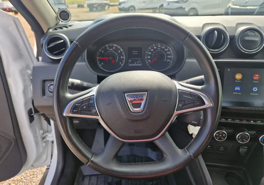 Vue centrée sur le volant noir du Dacia Duster blanc 2022, avec tableau de bord et écran tactile visibles.