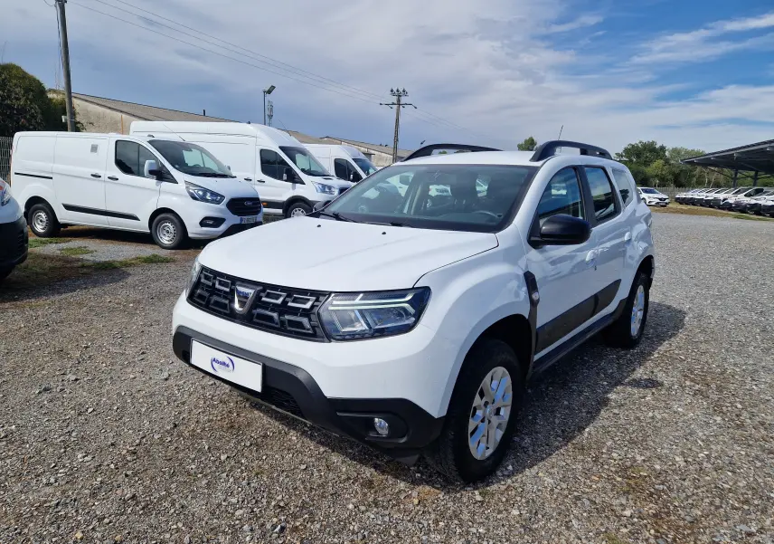 Dacia Duster blanc en 3/4 avant droit, avec barres de toit noires et protections latérales visibles.