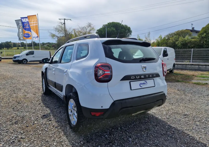 Vue 3/4 arrière droite d'un Dacia Duster blanc 2022 avec barres de toit et protections noires sur un terrain gravillonné.