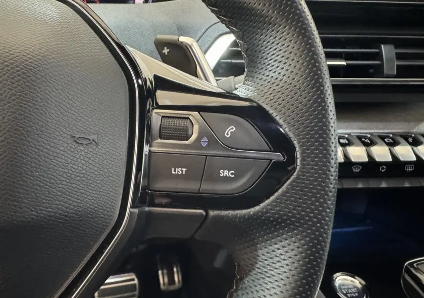 Gros plan sur le volant cuir perforé de la Peugeot 3008 GT, avec commandes audio et tableau de bord noir.