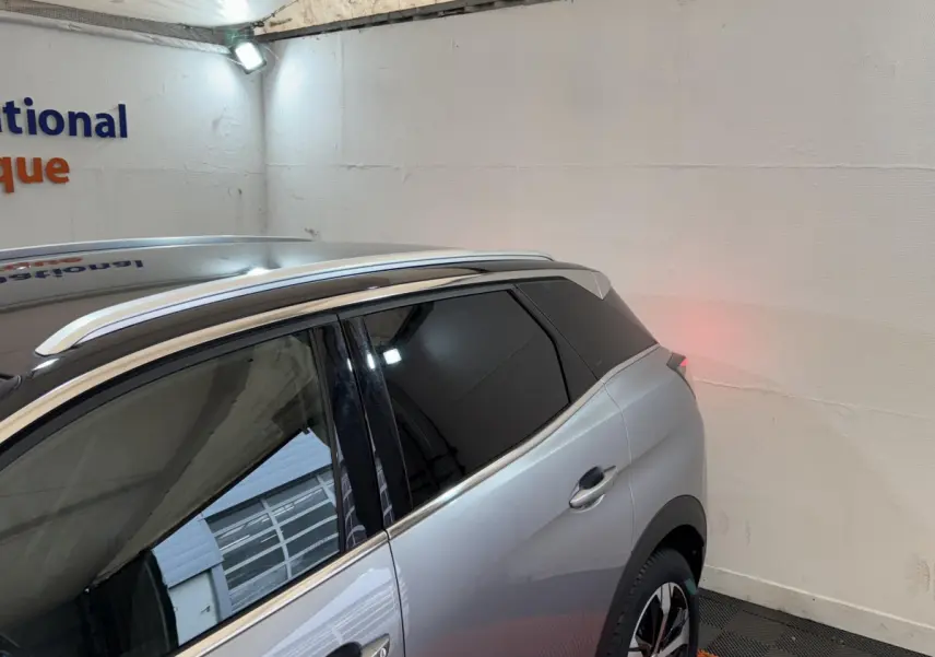 Vue 3/4 arrière droit du Peugeot 3008 GT gris clair avec vitres teintées et barres de toit chromées dans un garage.