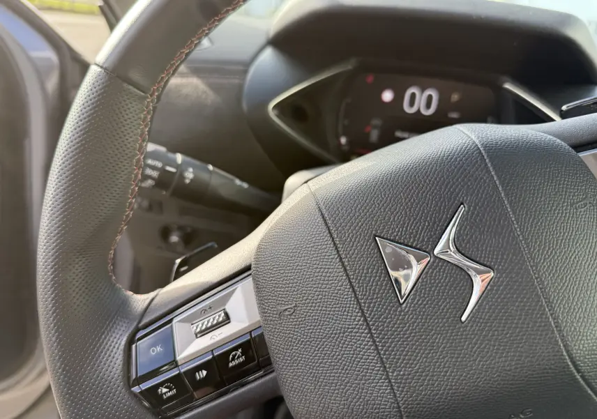 Gros plan sur le volant en cuir noir avec logo DS, tableau de bord numérique en arrière-plan flou.