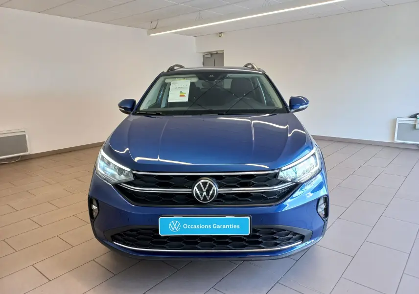 Vue avant d'un Volkswagen Taigo bleu récif 2025 en showroom, avec calandre noire et phares allumés.