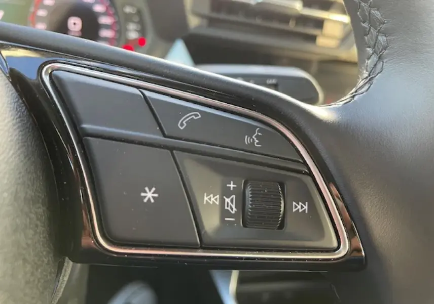 Gros plan sur les commandes du volant en cuir noir de l'Audi A3 IV Berline 30 TFSI MHEV argent métallisé, avec tableau de bord flou.