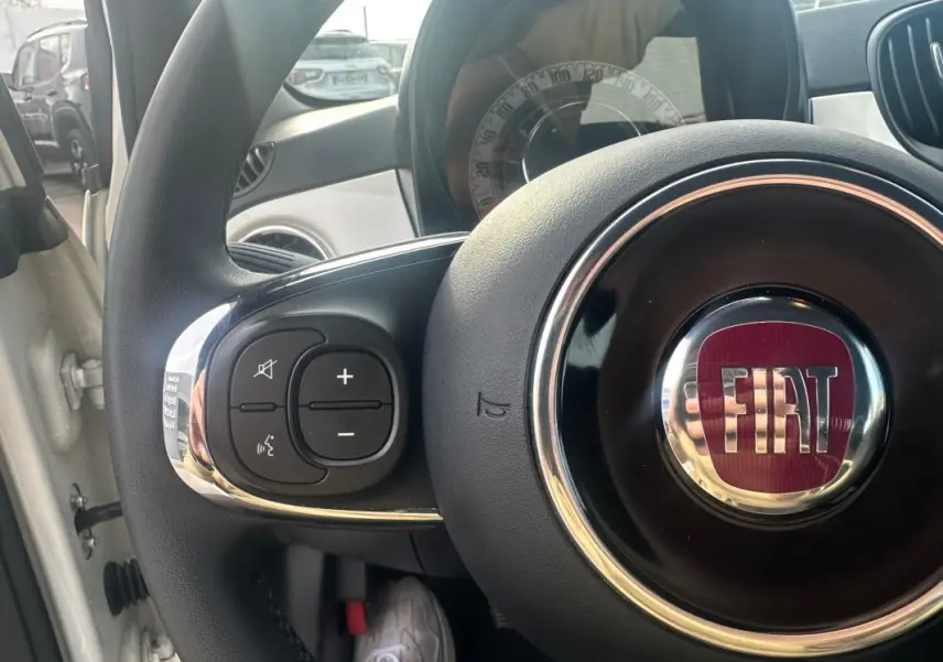 Gros plan sur le volant noir du Fiat 500 blanc 2023, avec logo FIAT rouge et commandes audio intégrées à gauche.