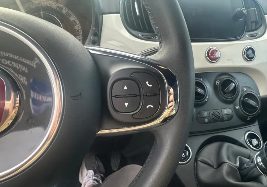 Vue intérieure centrée sur le volant de la Fiat 500 blanche 2023, avec commandes au volant et tableau de bord clair.