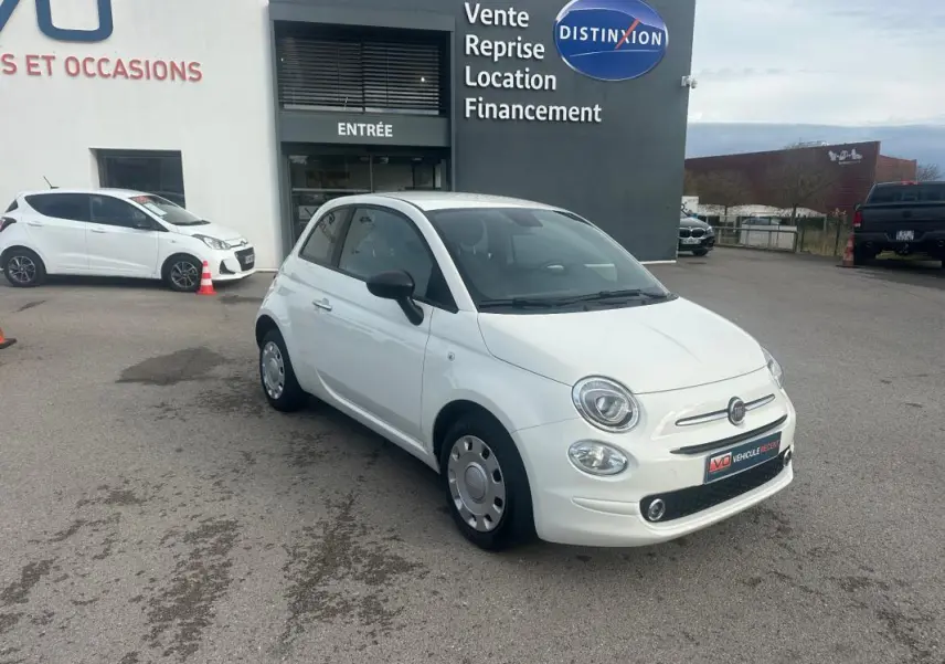 Vue 3/4 avant d'une FIAT 500 blanche de 2023, version hybride, avec rétroviseurs noirs, stationnée devant un bâtiment commercial.