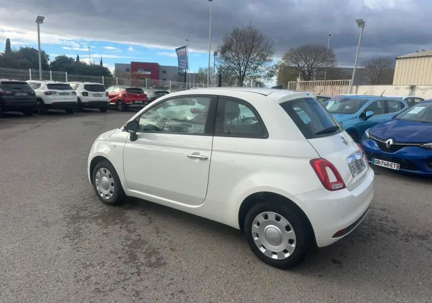 Vue 3/4 arrière droite d'une Fiat 500 blanche 2023, compacte et arrondie, stationnée en extérieur sur un parking.