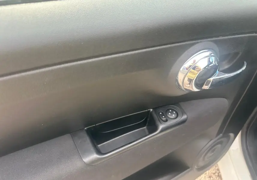Vue rapprochée de la poignée intérieure et des commandes de vitre côté gauche d'une FIAT 500 blanche 2023.