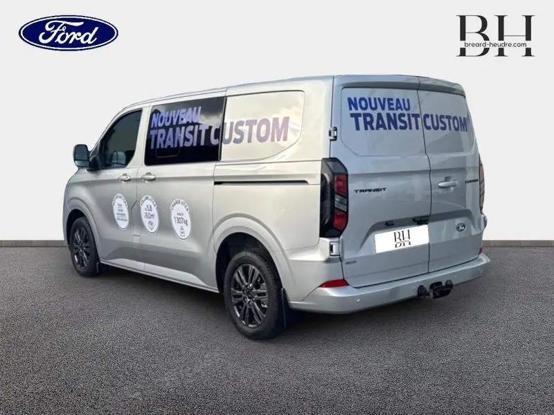 Vue 3/4 arrière droite du Ford Transit Custom gris lunaire métallisé avec marquages publicitaires et crochet d'attelage.