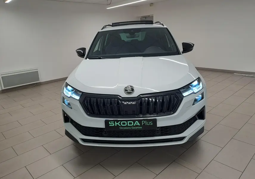 Vue frontale d’un Skoda Karoq Blanc Lune 2025 avec phares LED allumés et calandre noire Sportline.