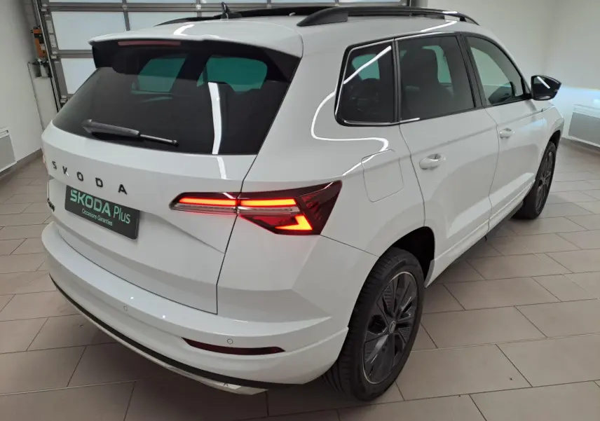 Vue 3/4 arrière droite d'un Skoda Karoq blanc lune 2025 avec feux arrière allumés et jantes noires sportline.