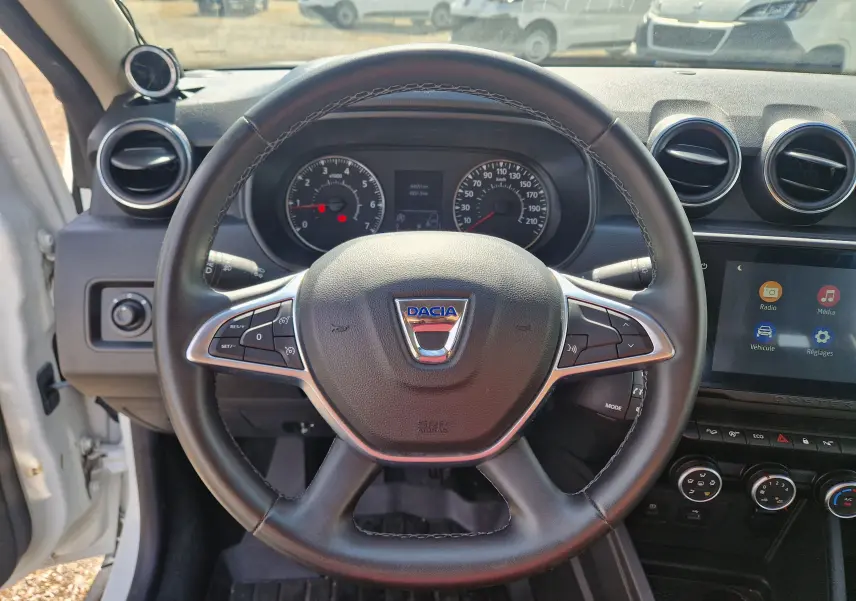 Vue intérieure centrée sur le volant noir du Dacia Duster blanc 2023 avec tableau de bord et écran tactile visible.