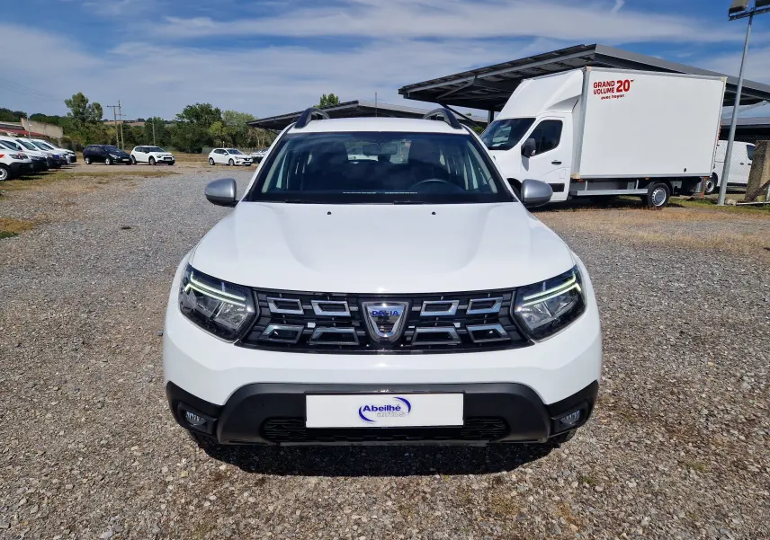 Vue frontale d'un Dacia Duster 4x4 blanc 2023 avec calandre noire et feux LED allumés sur un parking extérieur.