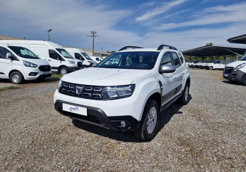 Dacia Duster blanc 2023 en 3/4 avant droit, SUV 4x4 avec barres de toit et jantes alliage visibles.