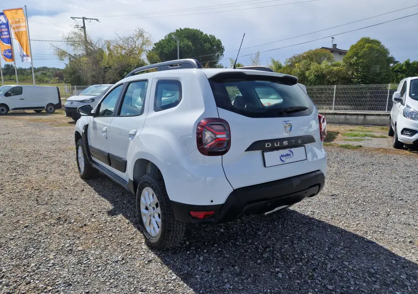 Vue 3/4 arrière droite du Dacia Duster blanc 4x4 2023, avec barres de toit et jantes alliage visibles.