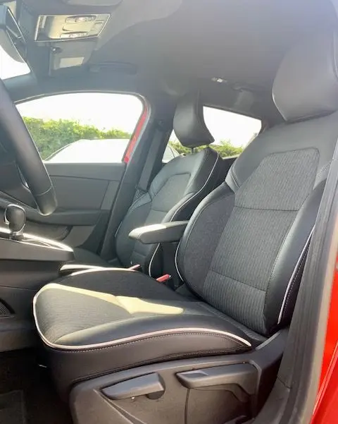 Vue intérieure côté conducteur de la Renault Clio V rouge 2021, mettant en valeur les sièges tissu et l'accoudoir central.