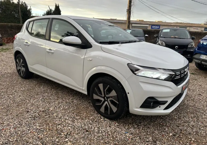 Dacia Sandero TCe 90 blanc vue 3/4 avant droit sur parking avec phares allumés et jantes stylisées.