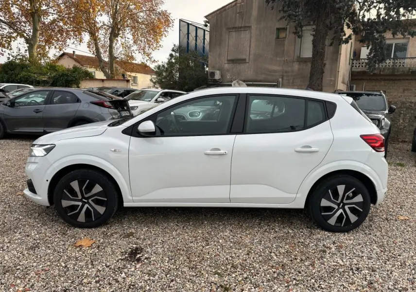 Profil côté gauche d'une Dacia Sandero blanche 2024 garée sur un sol gravillonné, avec jantes noires et grises.