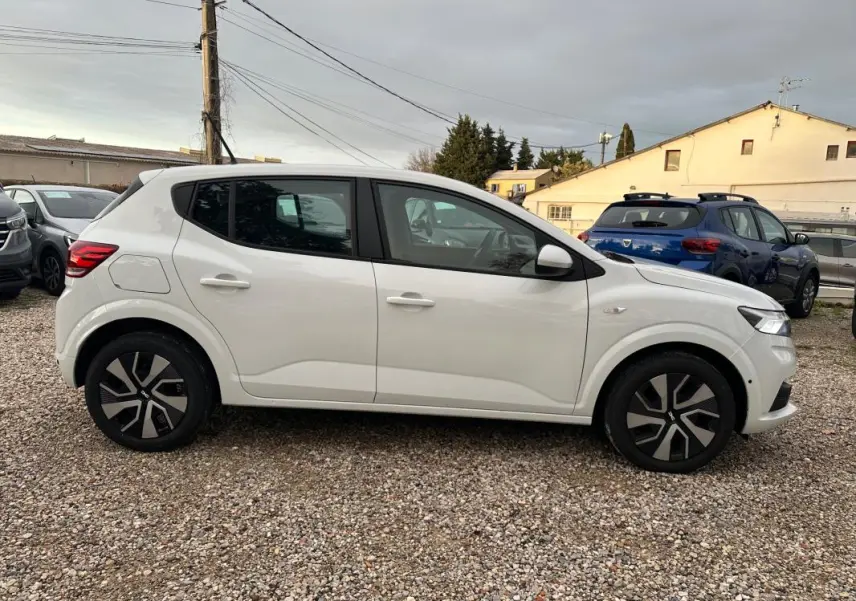 Vue de profil côté gauche d'une Dacia Sandero blanche 2024 garée sur un sol gravillonné, avec jantes noires et grises.