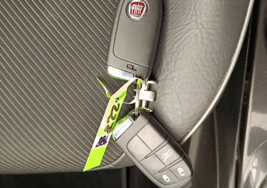 Clé FIAT posée sur siège tissu noir à coutures blanches, vue en plongée côté passager avant.