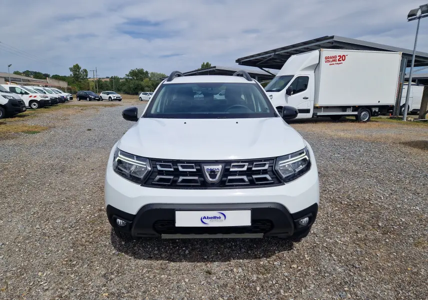 Vue frontale d'un Dacia Duster blanc 4x4 2022 avec calandre noire et phares LED allumés sur un terrain gravillonné.