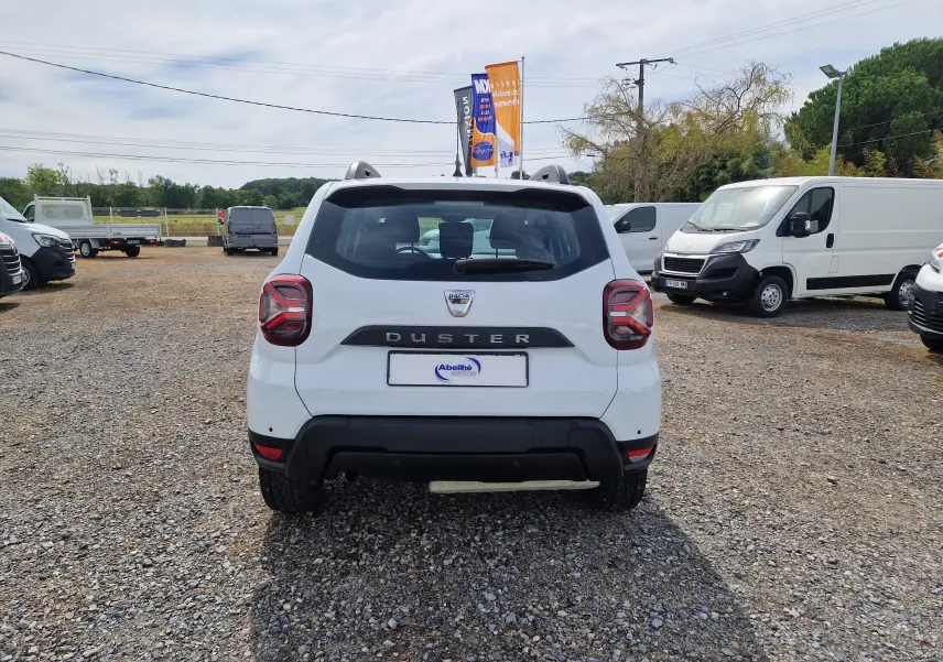 Vue arrière d'un Dacia Duster blanc 2022, avec logo et nom du modèle bien visibles sur un terrain gravillonné.