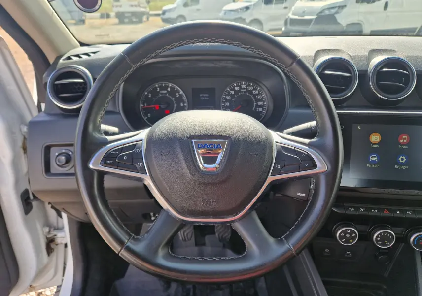 Vue intérieure centrée sur le volant cuir du Dacia Duster blanc 2023, avec tableau de bord et écran tactile visibles.