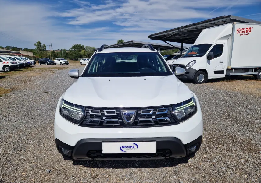Vue avant d'un Dacia Duster blanc 2023, avec calandre noire et feux à LED allumés, stationné sur un sol gravillonné.