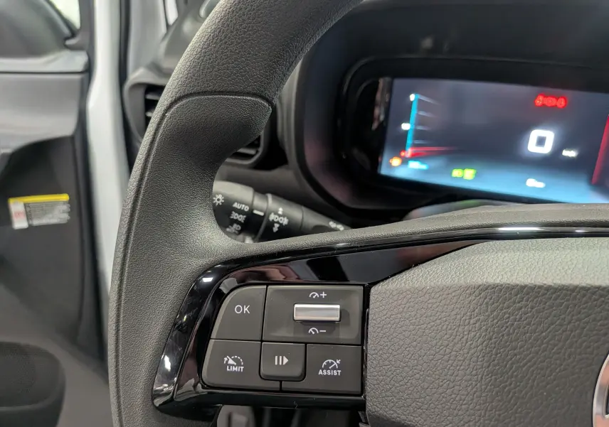 Gros plan sur le volant noir du Citroën Berlingo 2025 blanc, avec commandes de régulateur et tableau de bord numérique flou.