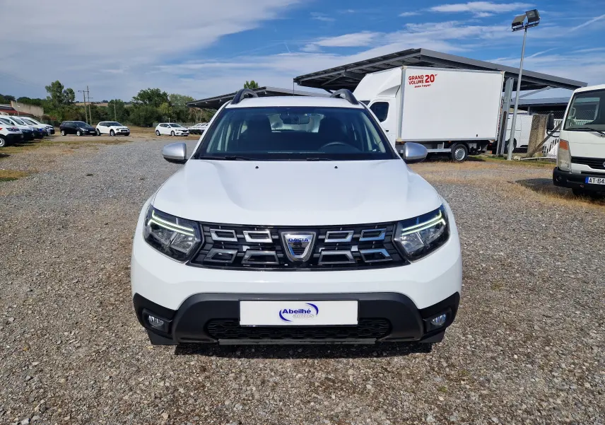 Vue frontale d'un Dacia Duster blanc 2023 avec calandre noire et feux LED allumés sur un terrain gravillonné.
